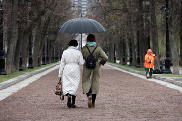 Two women with one umbrella walking on city street. Rainy weather in spring park
