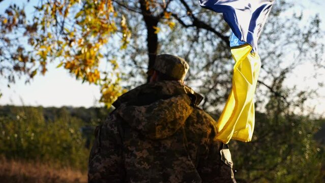 Young male military in uniform walks with waving flag of Ukraine at countryside. Soldier of ukrainian army going with blue-yellow banner outdoor. Concept of victory against russian aggression