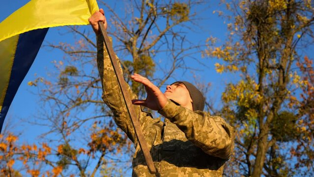Male ukrainian army soldier lifting blue-yellow banner in honor of victory against russian aggression. Young military in camouflage uniform waving flag of Ukraine at countryside. Invasion resistance
