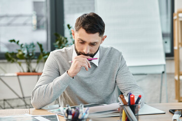 A man sits at a desk, engrossed in a piece of paper, deep in thought, surrounded by the hustle of a...