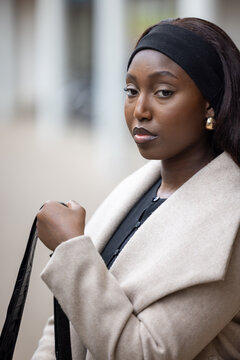 In this close-up, a young Black woman exudes urban elegance, clad in a stylish cream coat over a classic black outfit. Her headband and gold earrings add a touch of sophistication, while her gaze