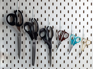 Different pair of scissors are hung on the pegboard. Scissors with different lengths of blades, with different shapes and ergonomics