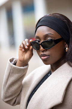 A fashion-forward Black woman stands on a city street, captured in the moment as she adjusts her sleek sunglasses. Her beige coat and headband epitomize urban chic, while her poised demeanor reflects
