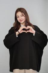 Smiling young woman making heart shape with hands, dressed in chic oversized attire. Symbolizes love and positive emotions.
