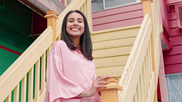 Young pretty smiling Indian woman, happy confident positive pretty girl from India wearing pink blouse standing outside countryside house on yellow stairs, looking at camera, portrait.
