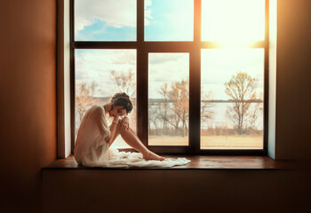 Fantasy young beautiful woman sitting on windowsill panoramic window. Romantic girl waiting for love, sky sunset sun rays divine light. vintage white dress. art style photo real person fashion model - obrazy, fototapety, plakaty