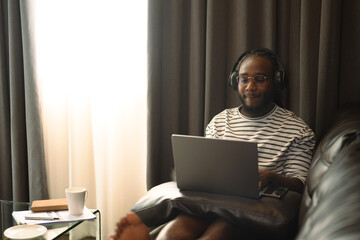 Young African American man freelancer in headphone working remotely on laptop at home