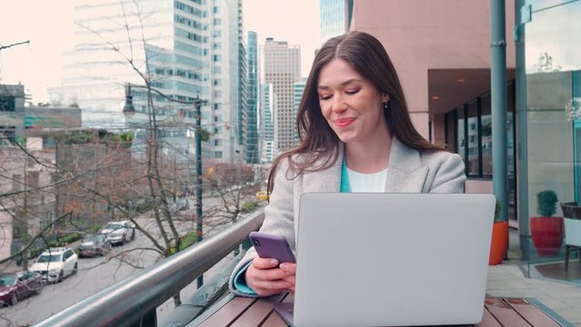 Young smiling happy business woman professional or student sitting outdoors on city street coworking terrace holding smartphone using mobile cell phone and laptop hybrid working or studying online.