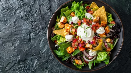 Fotobehang An enticing Mexican dish of chicken taco salad adorned with dollops of sour cream and salsa presented on a dark stone background from an aerial perspective © 2rogan