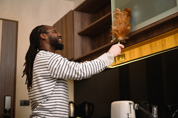 Happy young African man in casual clothes dusting shelf with a feather duster