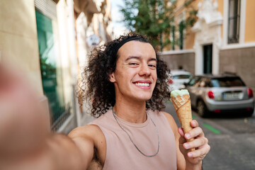 selfie of a cheerful man with long curly hair holding an ice cream cone in his hand in the city