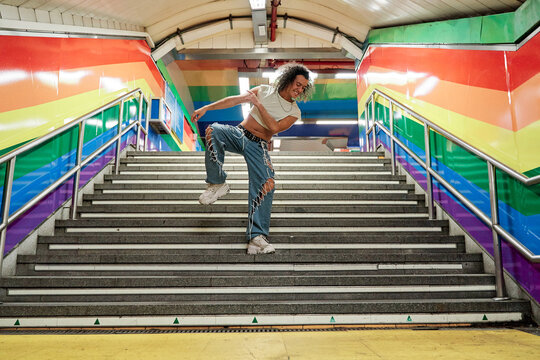 full view of a gay man dancing in the middle of the stairs of a subway station with the gay pride flag on the walls.