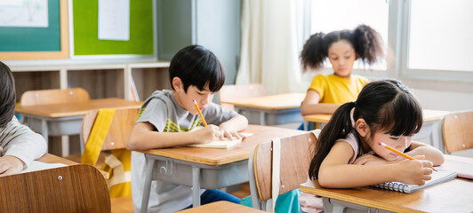 Portrait of little pupil writing at desk in classroom at the elementary school. Student girl study doing test in primary school. Children writing notes in classroom. Education knowledge concept