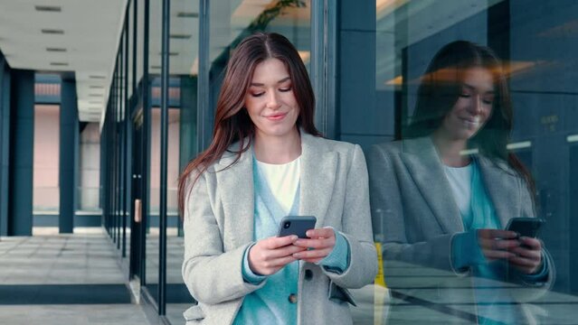 Young smiling happy woman, busy business lady standing outside office building on city street holding smartphone device using mobile cell phone technology, texting on cellphone.
