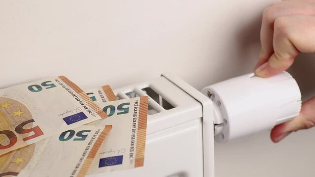 Amid escalating heating expenses, a hand cautiously turns the thermostat, slowly increasing. 50 euro bills lie on the radiator, highlighting the surge in costs.