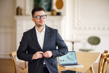 A poised and professional man buttoning his suit jacket stands by a work table with a laptop, in a...