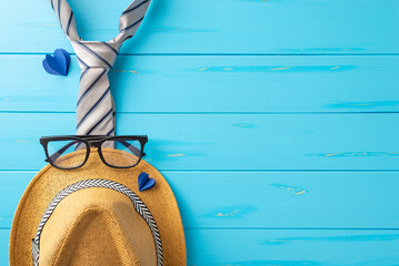 Celebrate Dad with a top view shot of a straw hat, sleek necktie, glasses, and paper hearts on a soft blue wooden desk backdrop. Perfect for Father's Day greetings or promotions