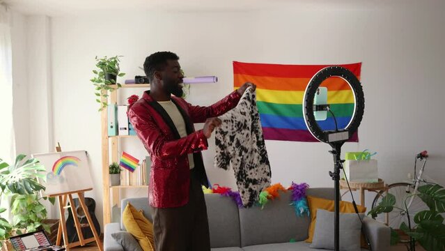 handsome african american blogger with sequin blazer showing clothes in front of camera recording online for his social networks, preparing outfit for gay pride party. share and display clothes online