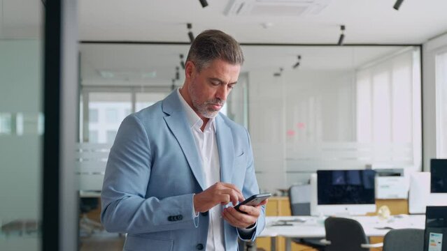Busy middle aged business man ceo investor standing in office using mobile cell phone. Mature businessman professional entrepreneur looking at smartphone at work making baking payment.