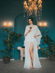 Fantasy woman hand holding glass of red wine. Sexy beauty face lady red lips pink make-up dark hair girl in luxury room. Long bare legs silver high heel shoes vintage silk white dress fairy art style 