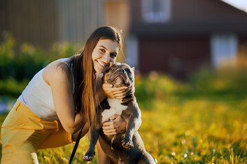 Trying to force dog to look into camera lens, young white woman takes photo with an American Bully in backyard of country house on summer evening.