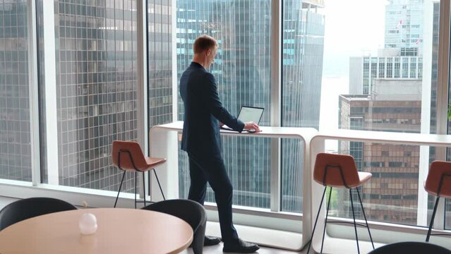 Busy professional business man employee, young male executive, businessman entrepreneur typing on laptop computer using phone working at office coworking standing near big skyscraper window. Full body