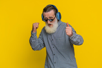 Excited senior stylish old man listening music via headphones and dancing disco party fooling around having fun expressive gesticulating hands. Happy elderly grandfather pensioner on yellow background - obrazy, fototapety, plakaty