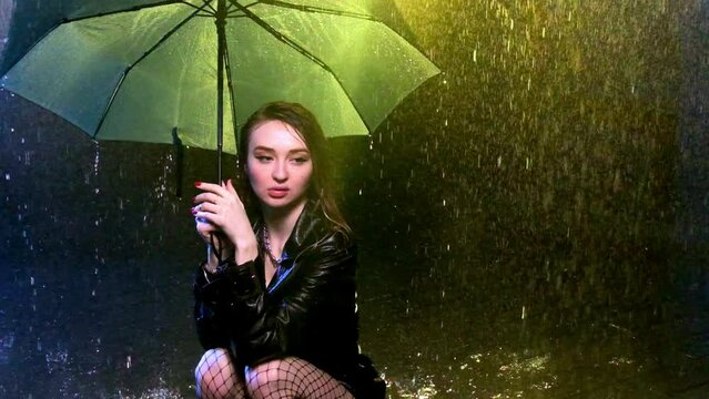 Woman wearing leather coat crouches under parasol in dimness of area. Streak of light accentuates umbrella and downpour