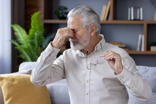 Tired gray-haired man sitting on the sofa at home, holding glasses in his hand and rubbing his eyes with his hand, resting - Powered by Adobe