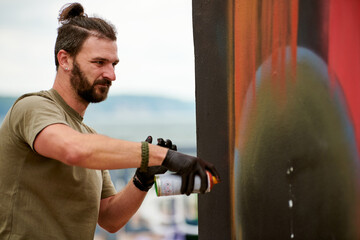 Attractive male artist is painting picture with paint spray can spraying it onto canvas at outdoor street exhibition, side view of man art maker