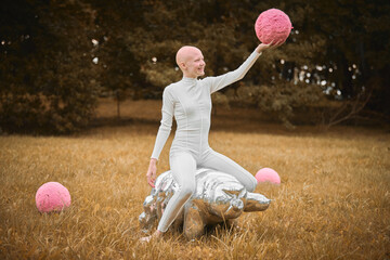 Young hairless girl with alopecia in white cloth sits on tardigrade figure and holds pink ball in hand on fall lawn park, surreal scene with bald teenage girl engages with symbolic elements - obrazy, fototapety, plakaty