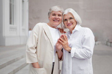 Portrait of beautiful smiling elderly senior women with gray hair dressed in stylish clothing, mature female models on city street against a bright building background - obrazy, fototapety, plakaty