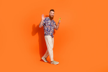 Full length photo of pretty friendly guy dressed print shirt chatting gadget walking waving hi isolated orange color background