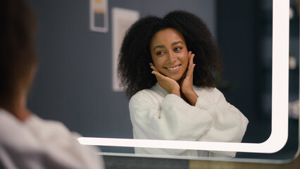 Happy smiling satisfied African American woman looking in mirror feeling joyful pampering in...