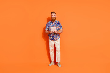 Full length photo of cheerful cool guy with red beard wear print shirt hold tablet browsing eshop isolated on orange color background