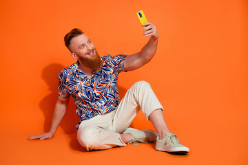 Full length photo of handsome guy wear print shirt white trousers sit making selfie on smartphone isolated on orange color background