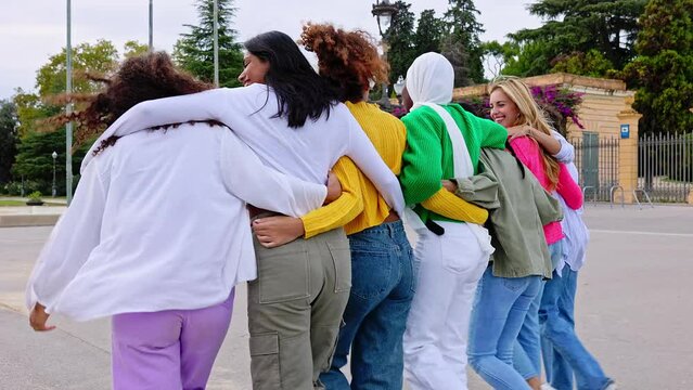 Rear view of young group of diverse women walking together embracing outdoors. Female friends hugging each other at city street. Friendship and female community concept.