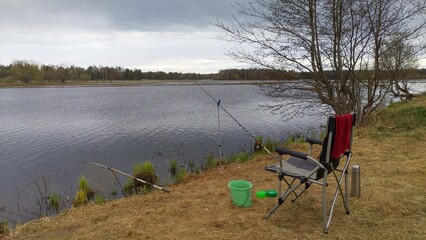 A folding fishing chair, landing net, a towel, bait boxes, a bucket with lure, a cage and feeder rod for bream fishing mounted on stands next to bushes on the river bank. Flood © Balser