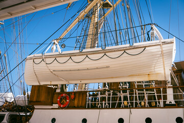 Lifeboat on sailing ship deck, clear blue sky background. Features include portholes and life ring....