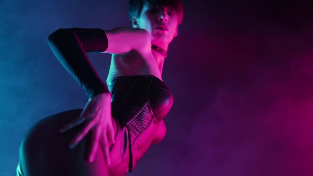 Sexy woman in bdsm leather corset performs dance moves in smoky neon light. Erotic hot fetish costume, dominatrix in dark studio. Dancer in night club.