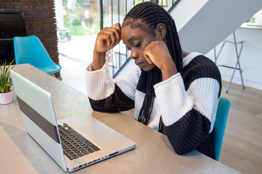 An African American woman with braided hair is deeply engrossed in her work, sitting at a modern desk. Her expression reflects concentration or concern as she stares at her laptop screen. She's