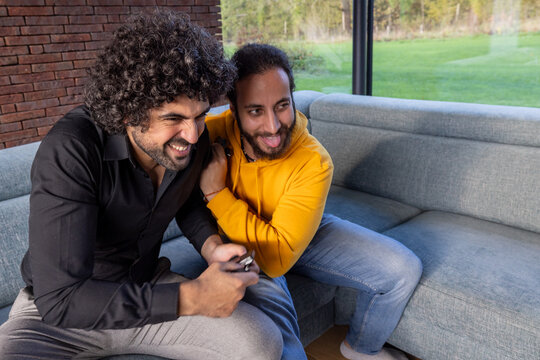 In a light-filled room with a view of the green outdoors, two friends share a sofa and a moment of pure joy. The man in the black shirt holds a smartphone, possibly sharing something humorous as they