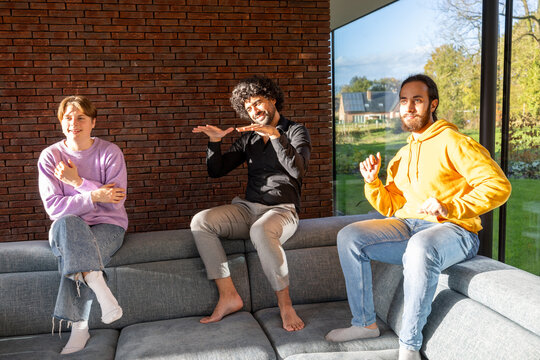 In a sunlit room with large windows and a brick wall, three barefoot individuals are seated on a grey sofa. They display animated expressions and gestures, suggesting a lively interaction. The person