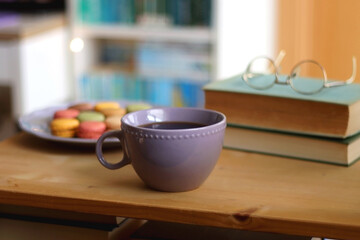 Fototapeta na wymiar Purple plate filled with pastel macarons, cup of tea or coffee, vintage books and reading glasses on the table. Colorful bookcase in the background. Selective focus.