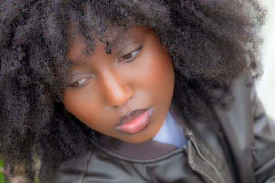 A close-up portrait captures a Black woman with voluminous natural afro hair and a subtle gaze. The soft focus on her features contrasts with her piercing eyes, expressing introspection. She's wearing