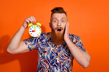 Photo of funky shocked guy dressed print shirt hand cheekbone rising clock isolated orange color background