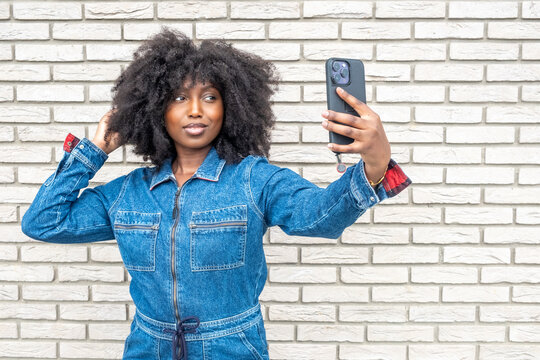 A young Black woman with fluffy, natural afro hair captures a selfie moment. Dressed in a full denim ensemble, she exudes a playful charm against the backdrop of a textured white brick wall