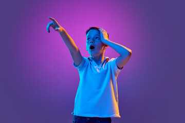 Wow. Surprised boy, in casual attire looking away and pointing to distance in neon light against purple gradient background. Concept of human emotions, childhood, education, fashion and style. Ad