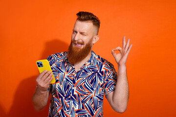 Photo of positive flirty guy dressed print shirt winking showing okey typing device empty space isolated orange color background