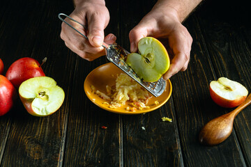 A grater in the hands of a chef for grating fresh apples for preparing dessert on the kitchen table.